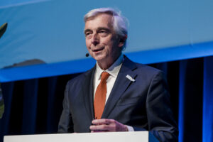 A person in a suit and orange tie stands at a podium, speaking into a microphone during a presentation.